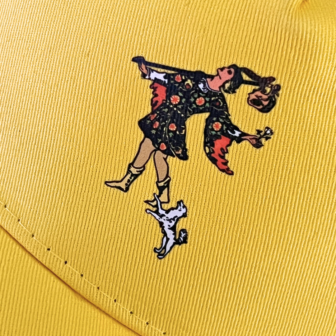 Close-up view of a yellow cap featuring an embroidered illustration of The Fool tarot card character alongside a small dog