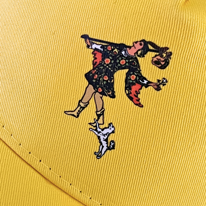 Close-up view of a yellow cap featuring an embroidered illustration of The Fool tarot card character alongside a small dog