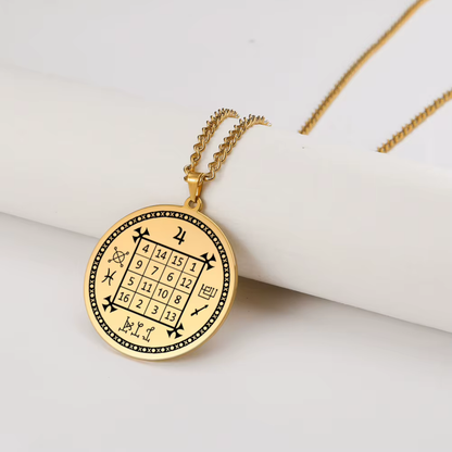 A gold-toned circular pendant necklace featuring an engraved magic square grid of numbers and mystical symbols, hanging from a matching chain on a white cylindrical prop against a white background.