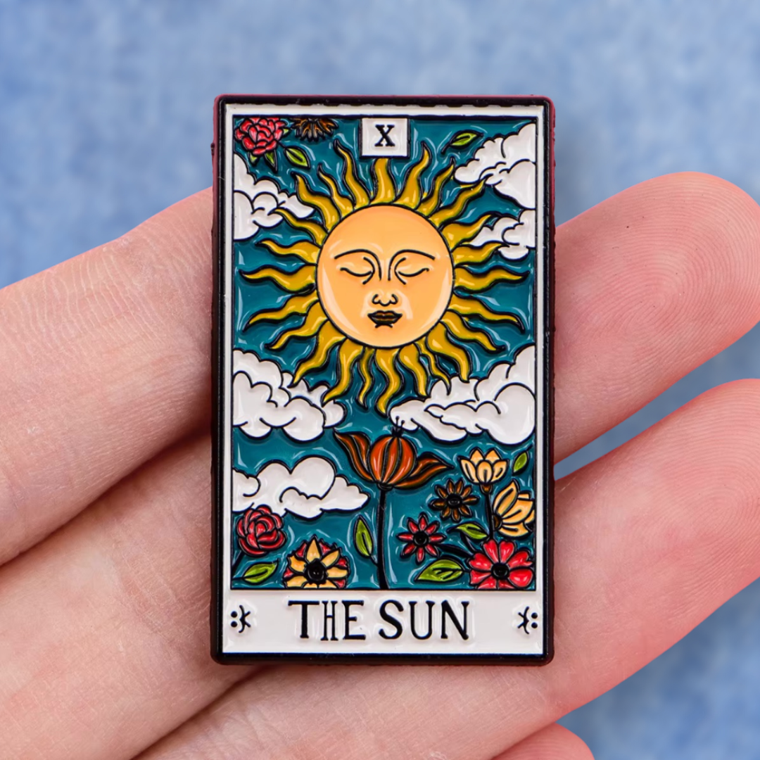 Hand holding The Sun tarot card enamel pin showing actual size scale and detailed view of the colorful vintage design against a blue jeans background