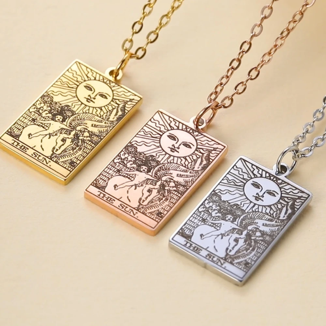 Three rectangular pendant necklaces on chains, each engraved with a tarot-style sun and figure design. The pendants are shown in gold, rose gold, and silver tones on a warm beige background.