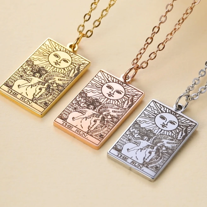 Three rectangular pendant necklaces on chains, each engraved with a tarot-style sun and figure design. The pendants are shown in gold, rose gold, and silver tones on a warm beige background.