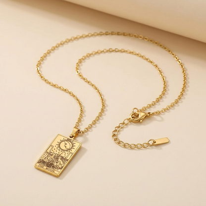 A gold-toned necklace with a rectangular tarot card pendant, displayed on a warm beige background.
