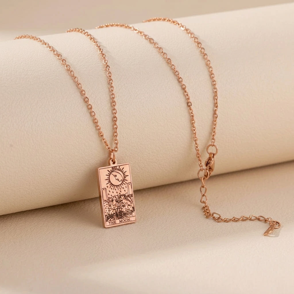 A rose gold necklace with a rectangular tarot card pendant featuring The Moon, displayed on a white cylindrical stand.