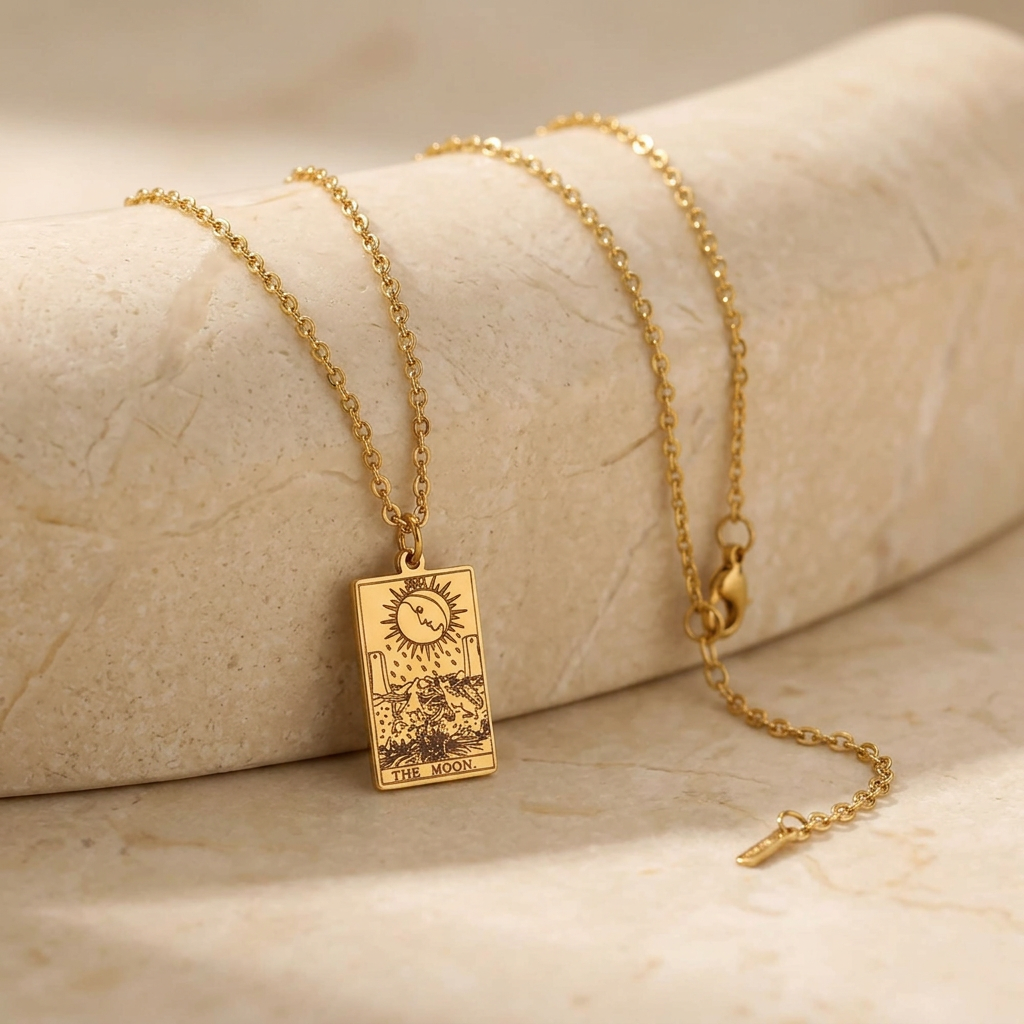 A gold-toned necklace with a rectangular tarot card pendant reading 'THE MOON,' draped over a light beige stone display on a matching stone surface.