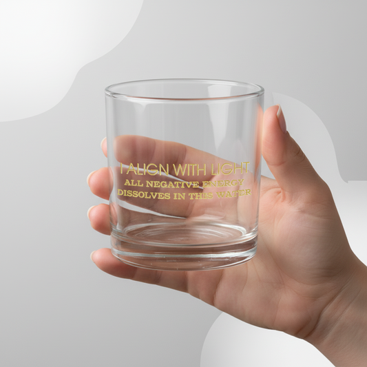 Clear glass with gold text that reads “I align with light. All negative energy dissolves in this water,” held in a hand against a light gray background.